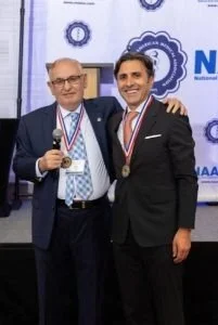 Two men in suits holding awards and medals, smiling at a conference or awards ceremony.
