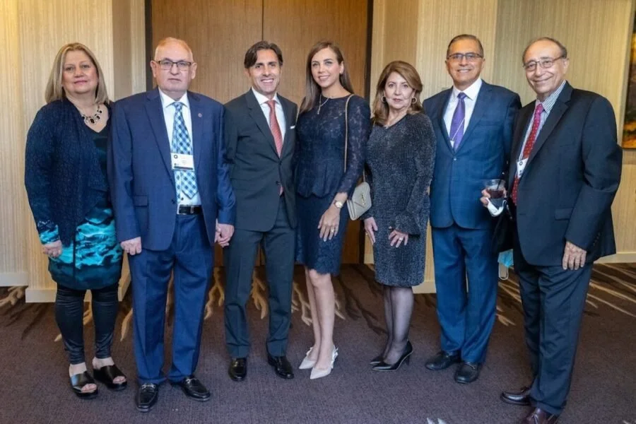 Group of seven professionally dressed people standing together indoors for a photo, some smiling, in a formal or business setting.