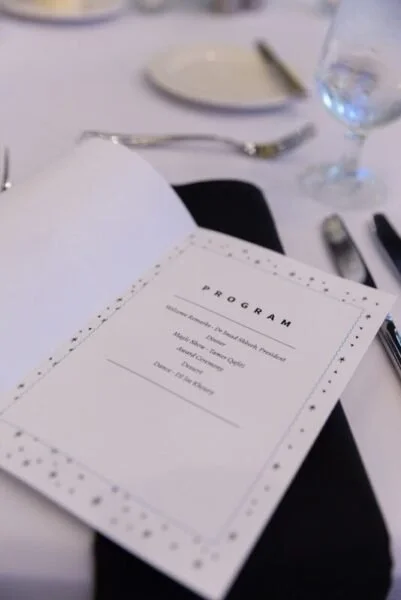 A wedding program on a dining table with a glass of water, a knife, fork, and a plate in the background.