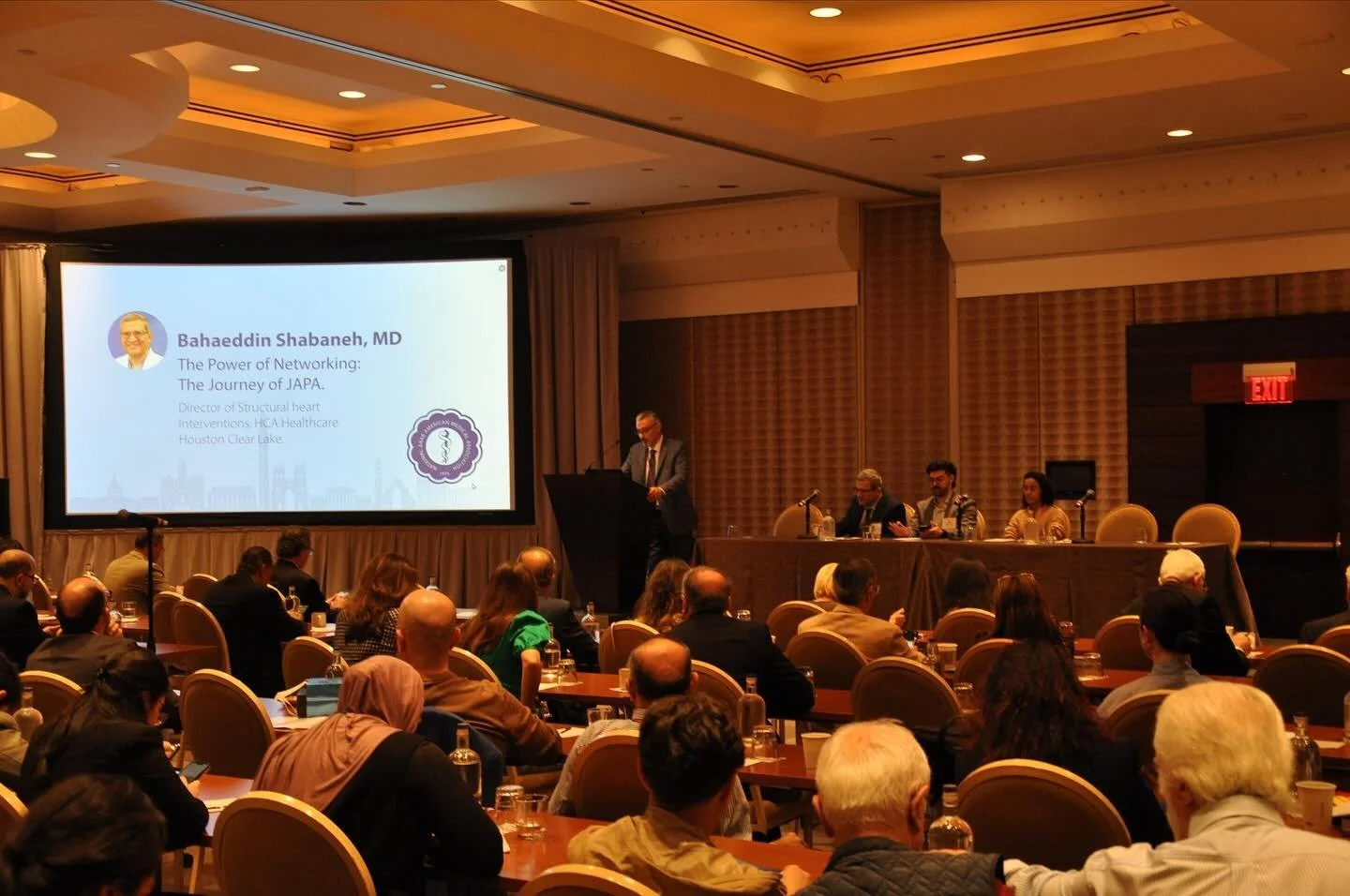 A speaker at a conference standing at a podium, with a large presentation screen displaying the name Bahaeddin Shabaneh, MD, and a panel of four people seated at a table on stage, in front of an audience in a conference room.