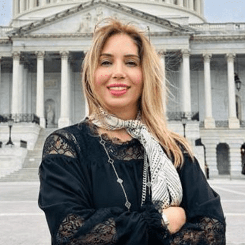 A woman standing in front of a large government building with pillars, smiling and wearing a black lace top and patterned scarf.