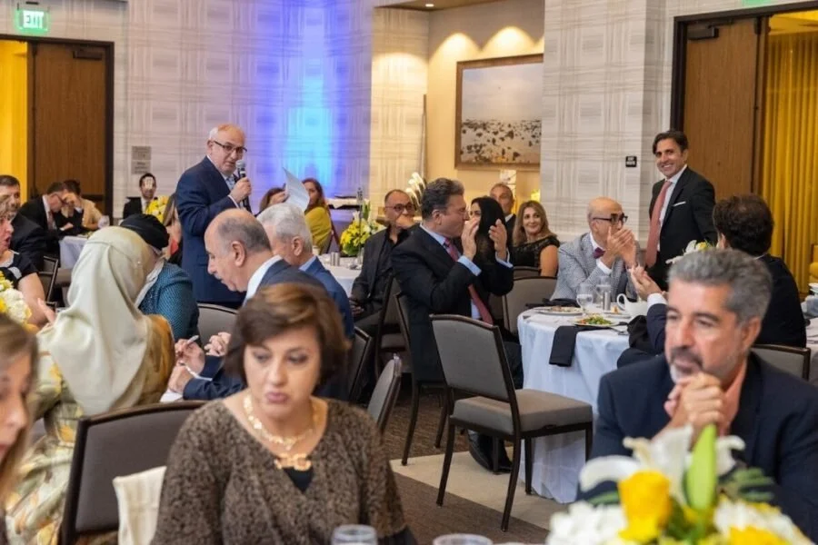 People attending a formal event or conference, with a man standing with a microphone, others are seated and clapping or listening.