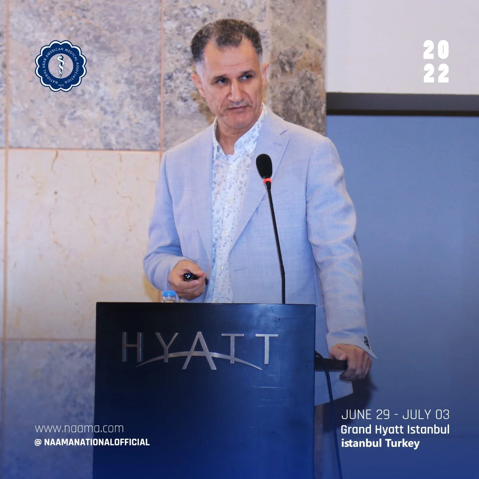 A man in a light gray suit giving a presentation at a conference in Istanbul, Turkey, held at the Grand Hyatt Istanbul hotel from June 29 to July 3, 2022. The background includes a marble wall and a blue podium with the Hyatt logo. There is a man hol