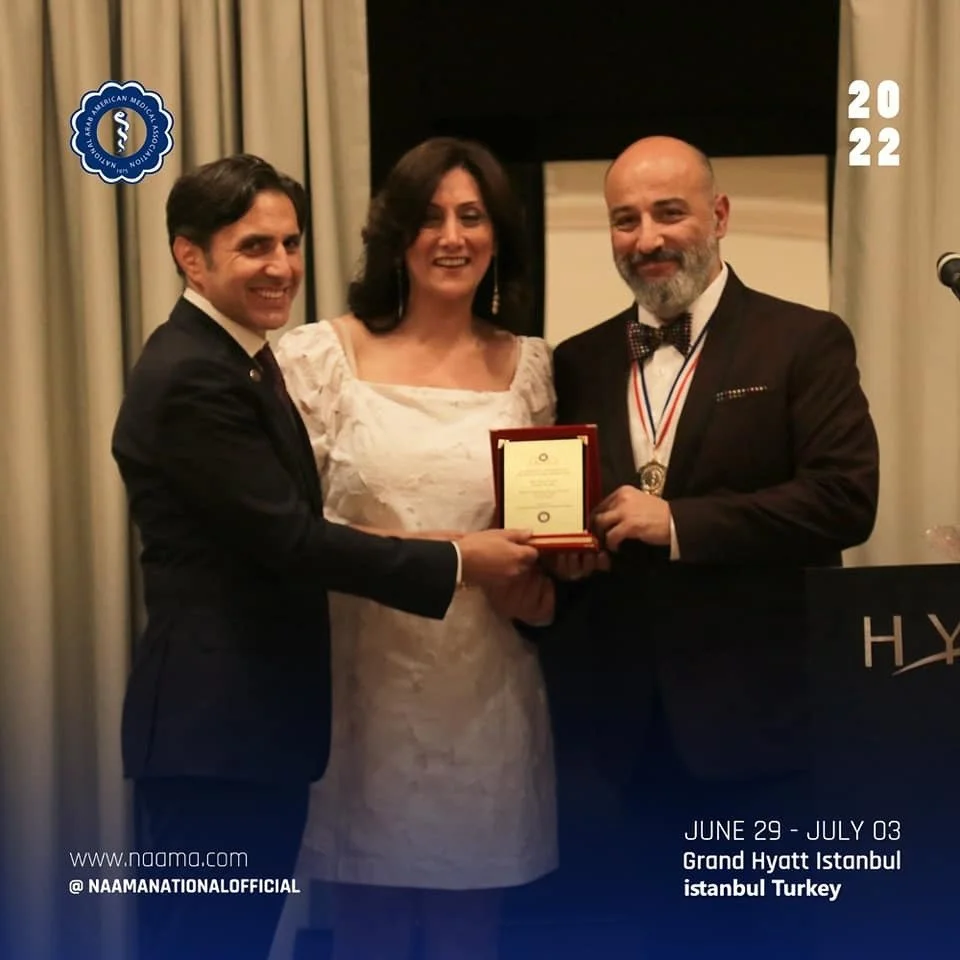 Three people standing together at an award ceremony, two men and one woman. One man is wearing a tuxedo with glasses and a medal, the woman is in a white dress, and the other man is in a dark suit. They are holding a plaque or certificate. The event 