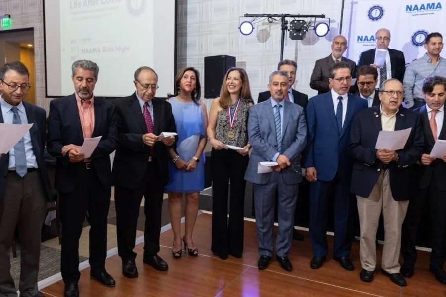 Group of professionally dressed people standing on a stage during an award ceremony, with some holding papers and medals, in a conference room with NAAMA branding.
