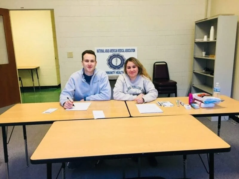 Two people sitting at a long table in a room, with a sign in the background that reads 'National Arab American Medical Association, Community Health'. The table has papers, pens, and a water bottle.