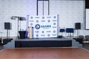 Empty stage with a NAAMA backdrop, two tall planters with plants, and chairs on either side, set up for a conference or event in a large room.