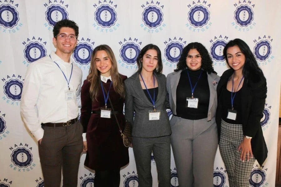 Five diverse young professionals, four women and one man, standing together smiling in front of a NAAMA NEXGEN branded backdrop at a conference.