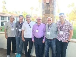 Six men standing outdoors, smiling for a group photo, some wearing conference badges around their necks, with a blurred background of trees and buildings.