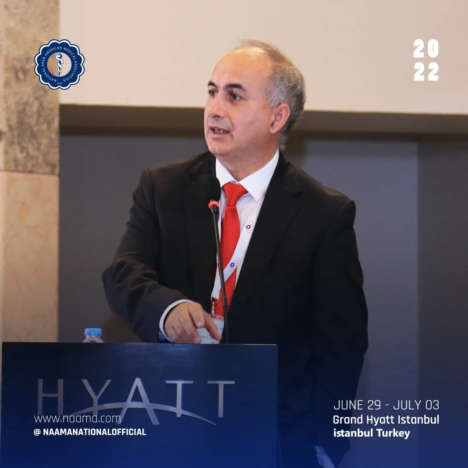 A man wearing a dark suit, white shirt, and red tie standing at a podium with a microphone. The podium displays the Naama logo and the event details: June 29 to July 3 at the Grand Hyatt Istanbul in Turkey. The background includes the logo of the Ara
