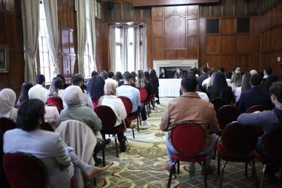 A large group of people attending a formal event or conference in a wood-paneled room with large windows, facing a panel of speakers at the front.