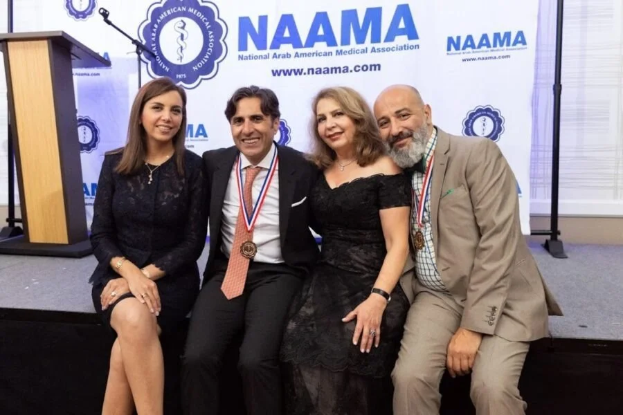 Four people sitting in front of a NAAMA (National Arab American Medical Association) backdrop, dressed in formal attire, with some wearing medals and ribbons around their necks, smiling at the camera.
