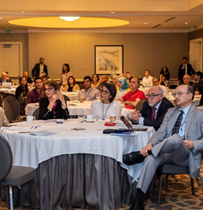 A group of people attending a conference or seminar, sitting at round tables with presentation materials, in a hotel conference room.