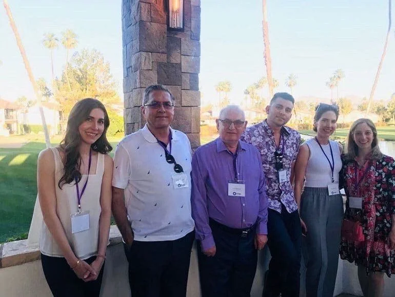 Six people standing outdoors at a social event, with palm trees and a lake in the background, posing for a group photo.