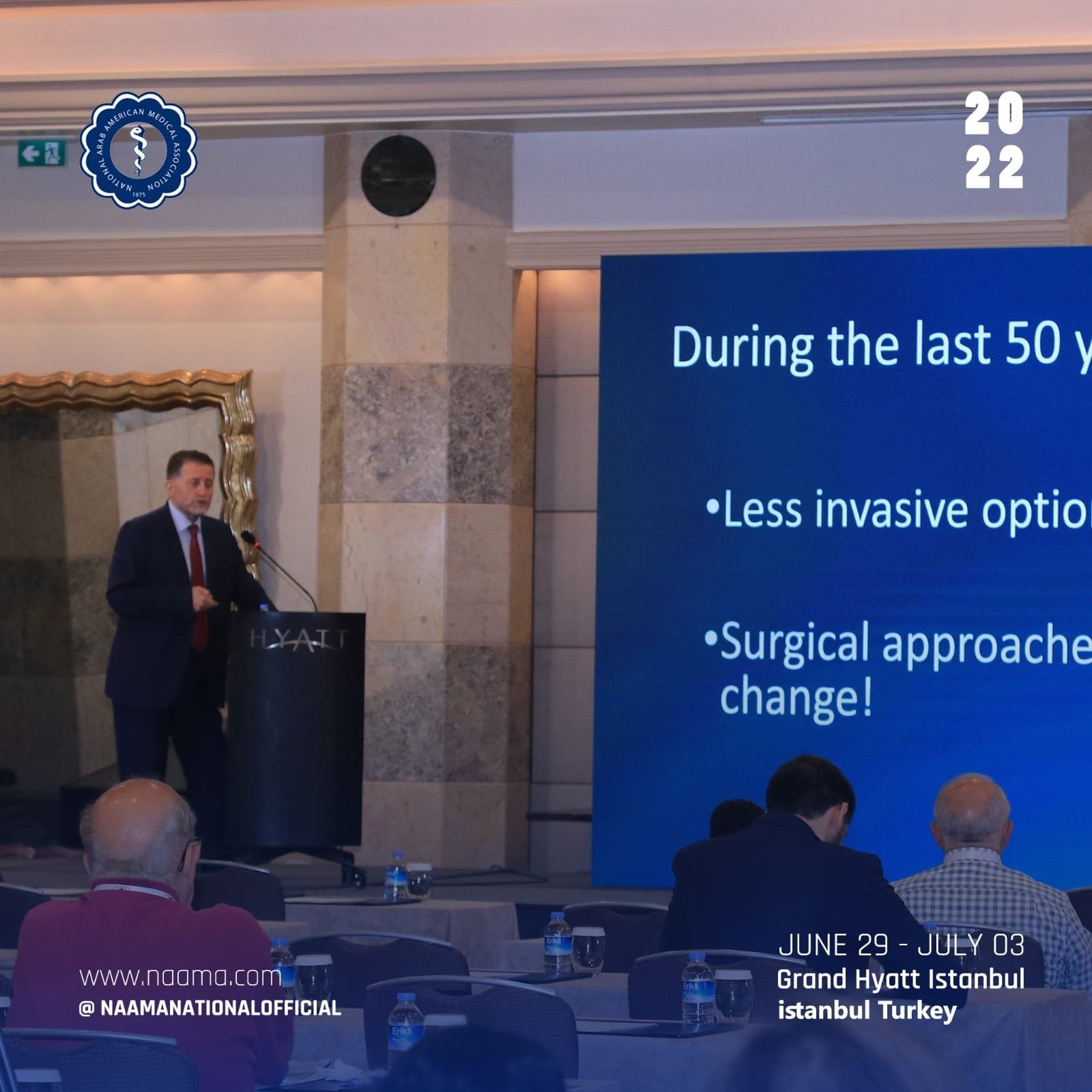 A man giving a presentation at a conference in a hotel ballroom. He is standing behind a podium with a microphone, and there is a large blue screen displaying text about medical options. There are several attendees seated at tables with water bottles