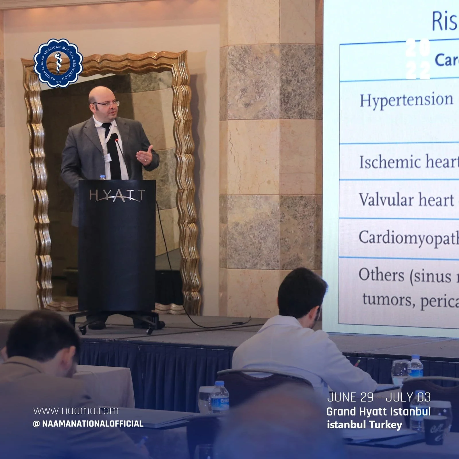 A man giving a presentation at a conference at the Grand Hyatt Istanbul, Turkey. He is standing at a podium branded with Hyatt and is speaking into a microphone. There are attendees seated at tables with water bottles, and a large screen displaying a