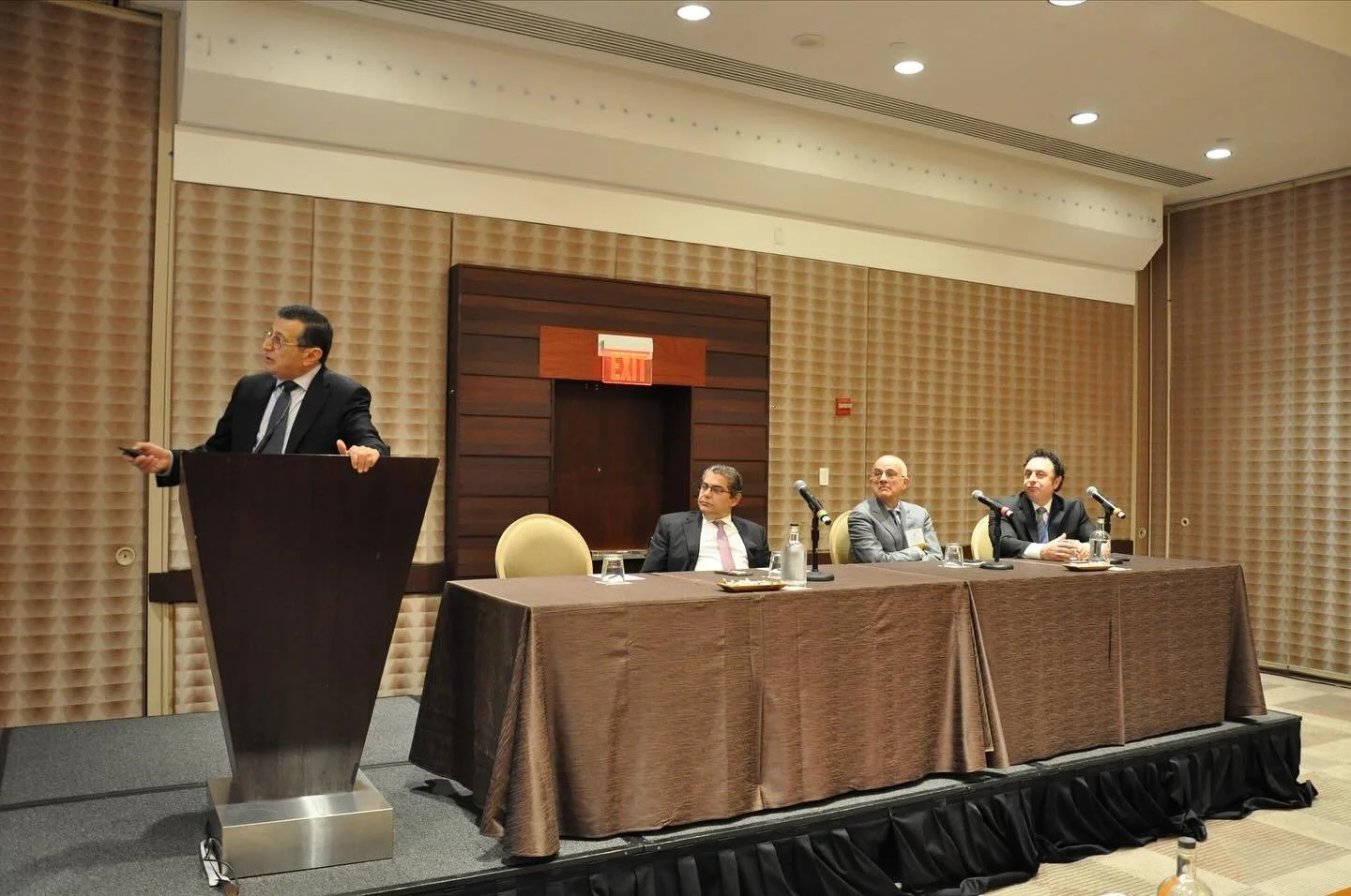 A man in a suit is standing at a podium giving a presentation while three other men in suits sit at a table with microphones in front of them during a conference or panel discussion.