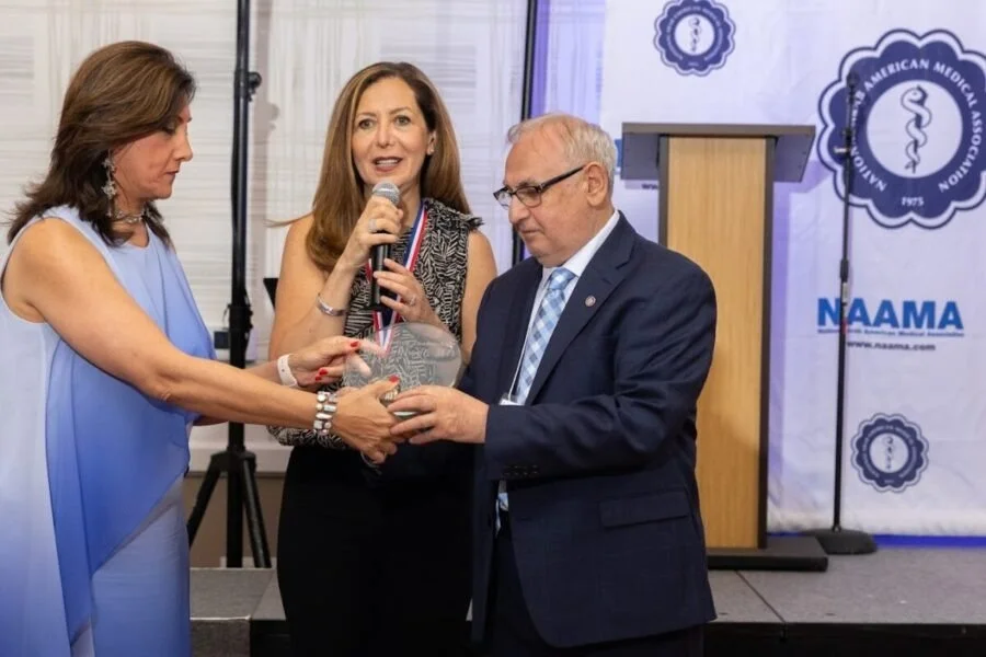 A woman holding a microphone presents an award to a man in a suit, while another woman in a blue dress looks on at an awards ceremony for the American Medical Association.