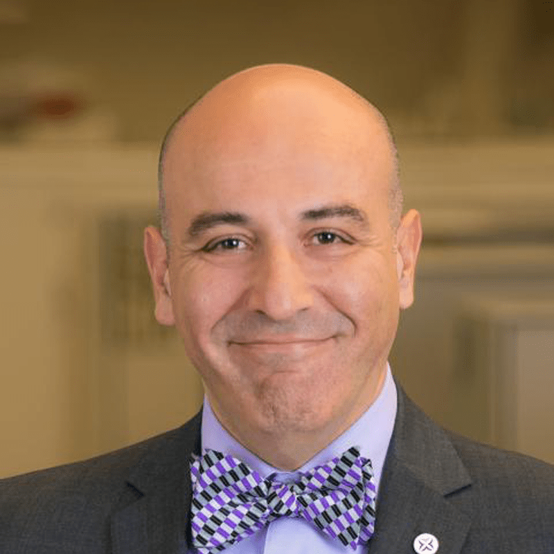 A man with a shaved head wearing a dark suit, purple shirt, and a checkered purple, black, and white bow tie, smiling in an indoor setting.