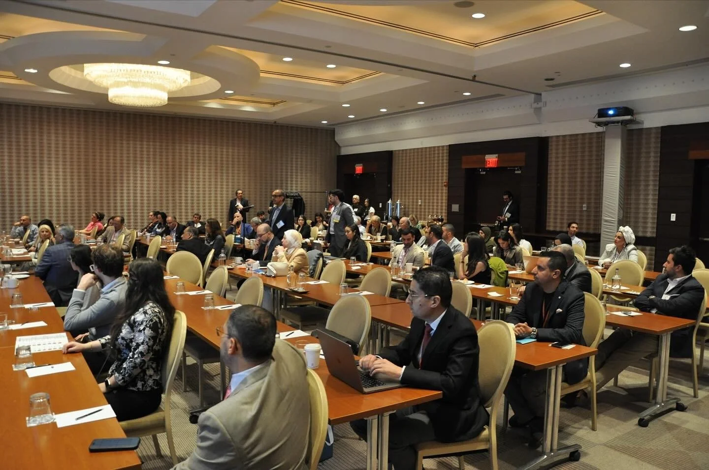 Conference room with attendees sitting at long tables, some using laptops, and a few standing and speaking, with a large chandelier on the ceiling.