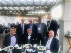 Seven men in formal attire gathered around a dining table at a banquet or event, some standing and some seated, with a large decorated venue in the background.