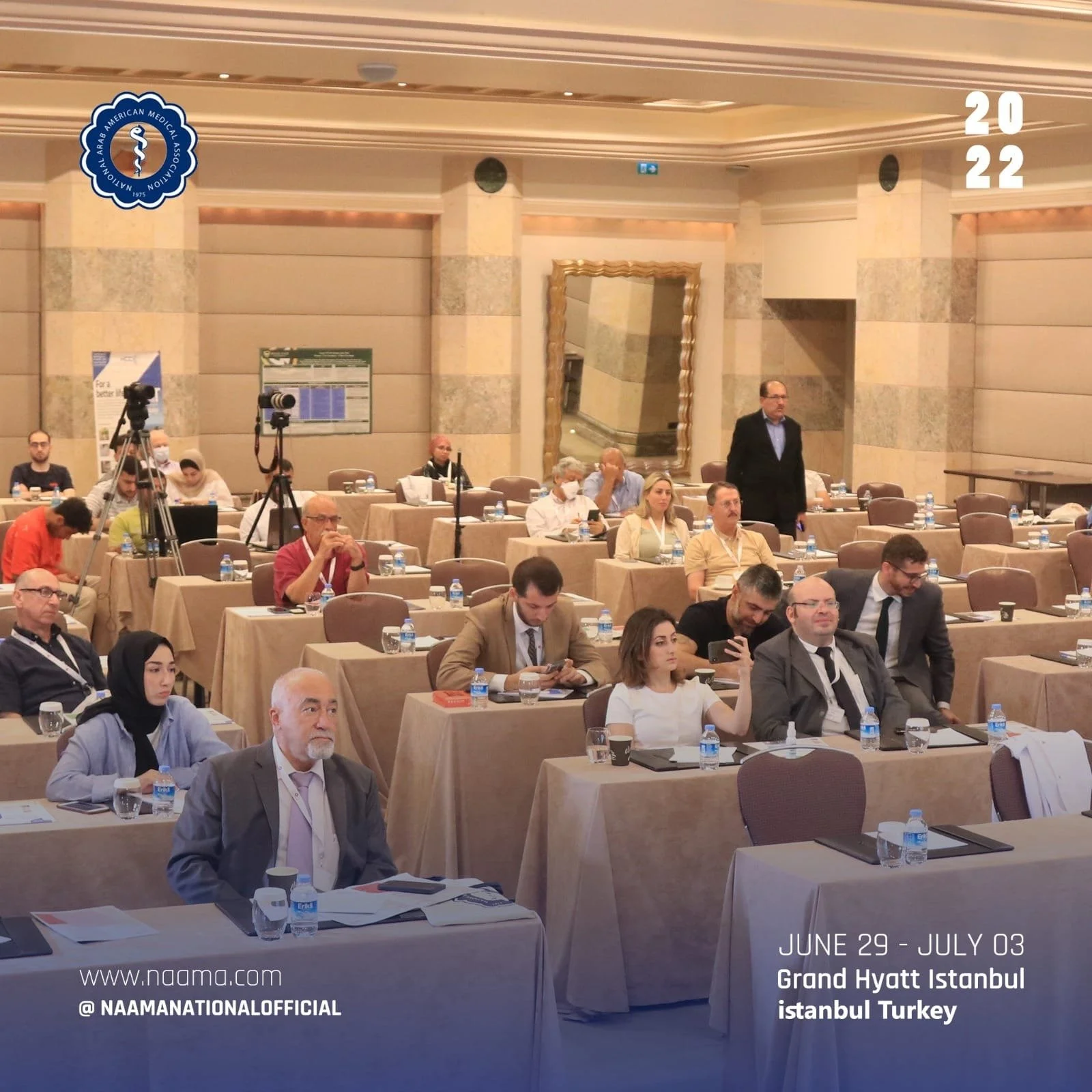 Conference attendees seated at tables in a hotel conference room at the Grand Hyatt Istanbul, Turkey, during the NAAM national official event from June 29 to July 3, 2022. Several people are seen using laptops and taking notes, with water bottles and