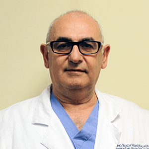 A middle-aged man wearing glasses and a white medical coat, standing against a plain light-colored background.