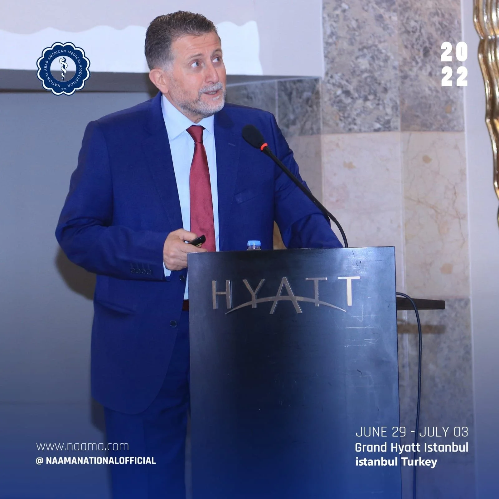 A man in a blue suit and red tie speaking at a podium with a microphone at the Grand Hyatt hotel in Istanbul, Turkey, during an event by the Arab American Medical Association, which took place from June 29 to July 3, 2022.