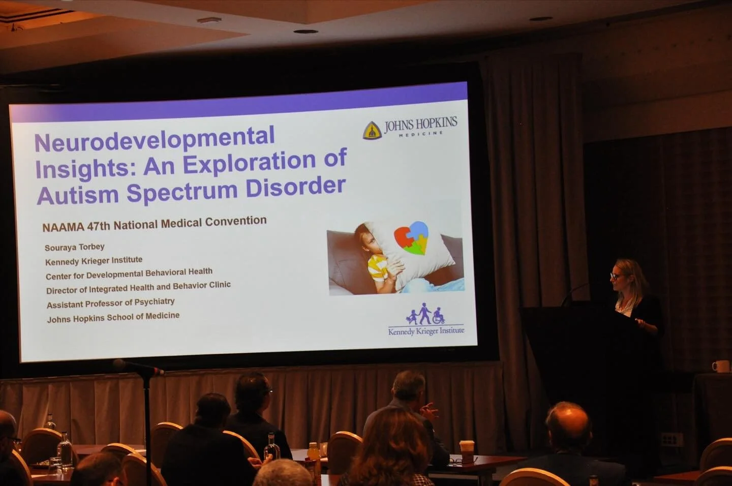 Presentation slide titled "Neurodevelopmental Insights: An Exploration of Autism Spectrum Disorder" at NAAMA 47th National Medical Convention, with a woman speaker at a podium to the right, audience members seated and listening.