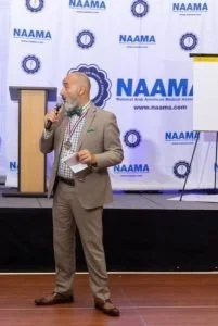 A man in a tan suit speaking into a microphone at a conference with a Nawsa banner in the background.