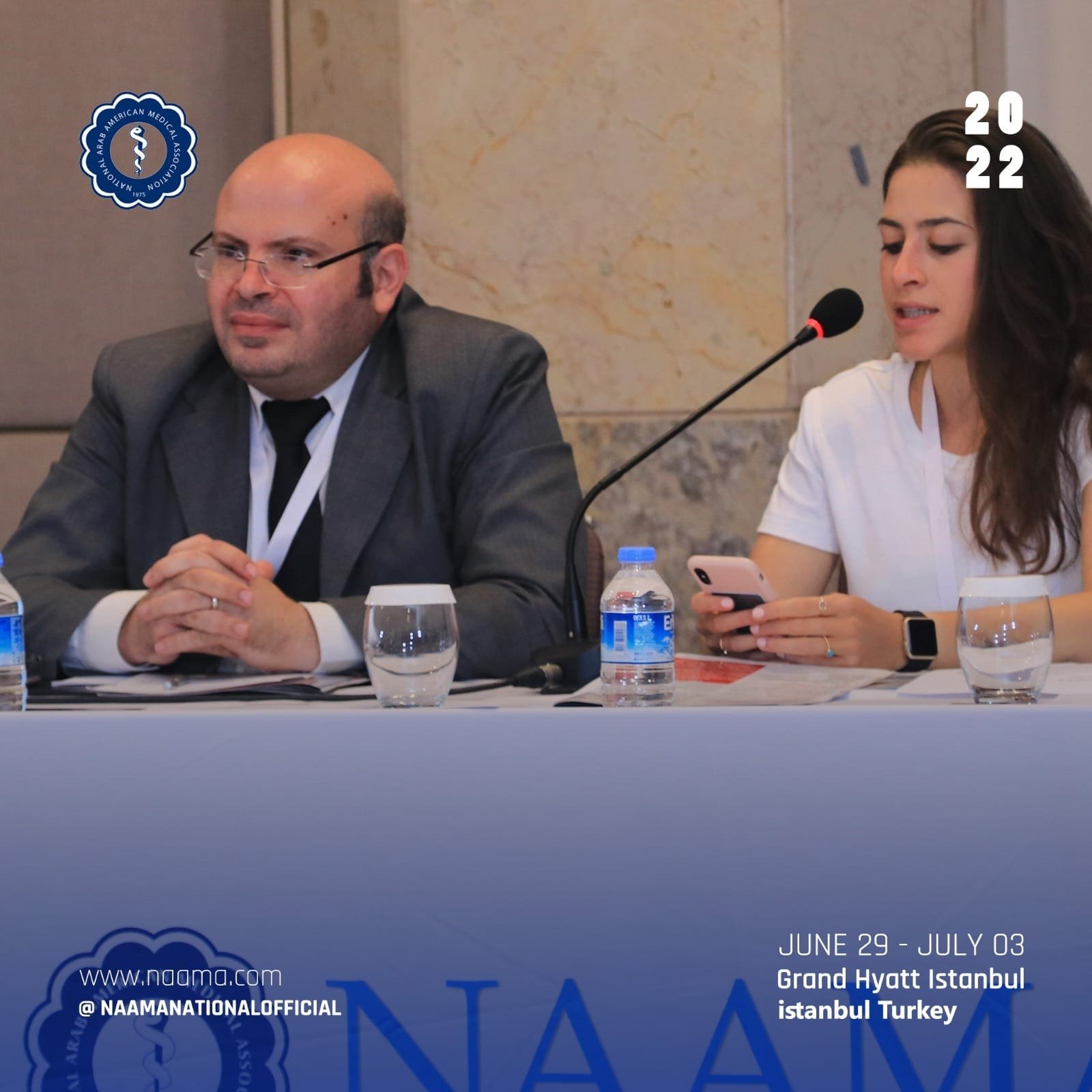 A man and a woman sitting at a conference table with a microphone, water bottles, and glasses in front of them. The man is bald, wearing glasses, a gray suit, white shirt, and black tie. The woman has long dark hair, is wearing a white shirt, and is 