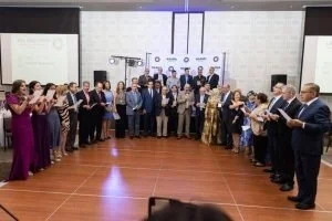 Group of formally dressed people standing in a circle at a party or celebration in an indoor venue with a large white wall and a social distancing reminder in the background.