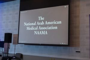 Projection screen displaying the text 'The National Arab American Medical Association NAMAA' at an indoor event.