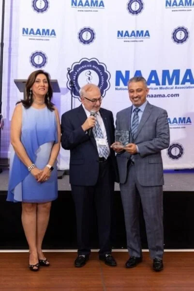 Three people on stage at an event with NAAMA banners, one man is receiving an award from another man, a woman stands beside them.
