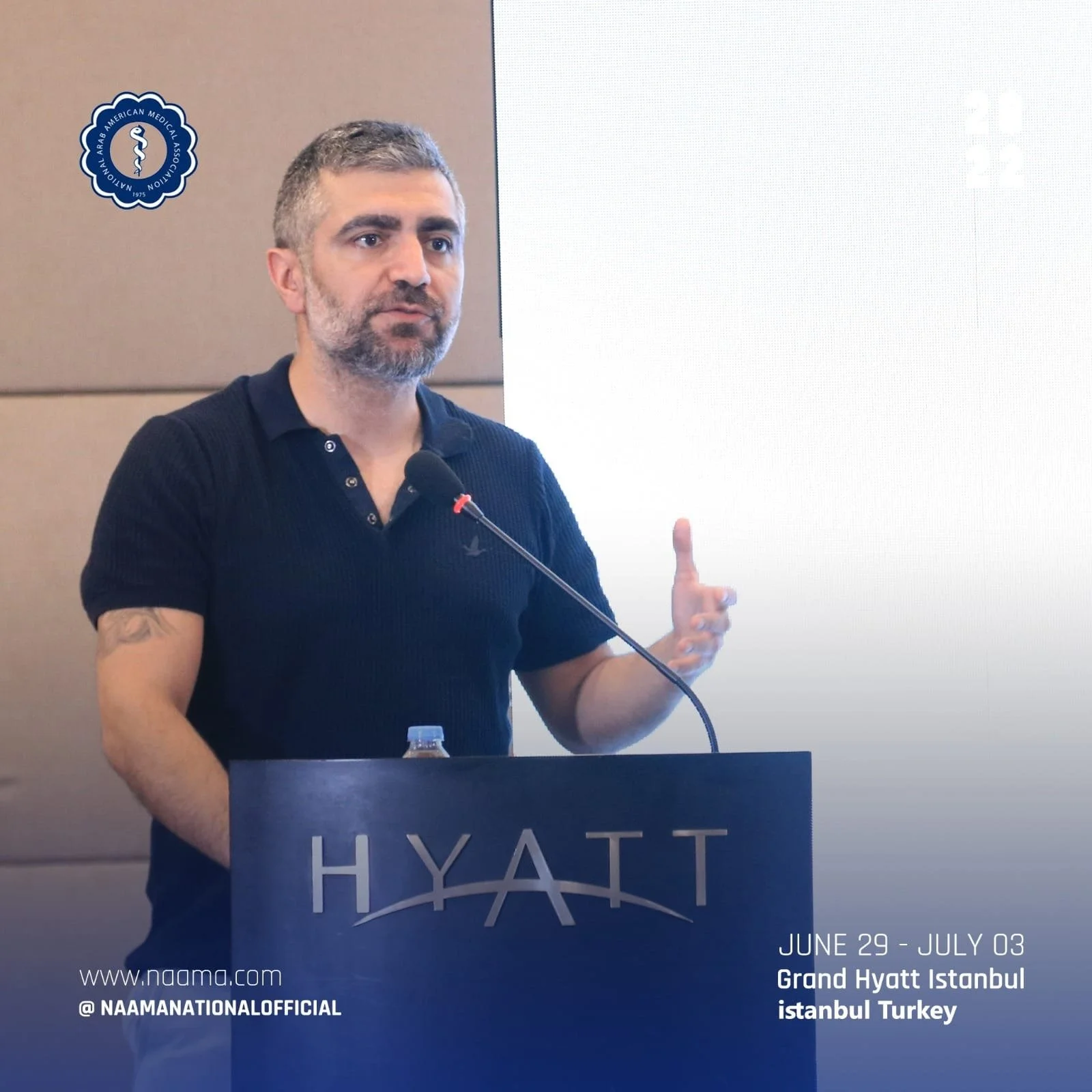 A man with gray hair and a beard, wearing a black polo shirt, speaking at a podium with a microphone. The podium has the Hyatt hotel logo, and a water bottle is visible on it. Behind him, there is a beige wall and a large white screen. The logo on th
