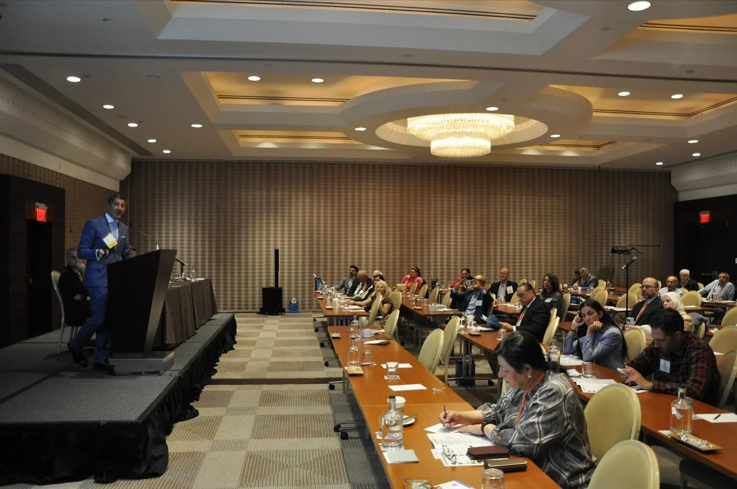 A man in a blue suit and glasses speaking at a podium during a conference in a large, well-lit hotel ballroom, with attendees seated at long tables taking notes and listening.