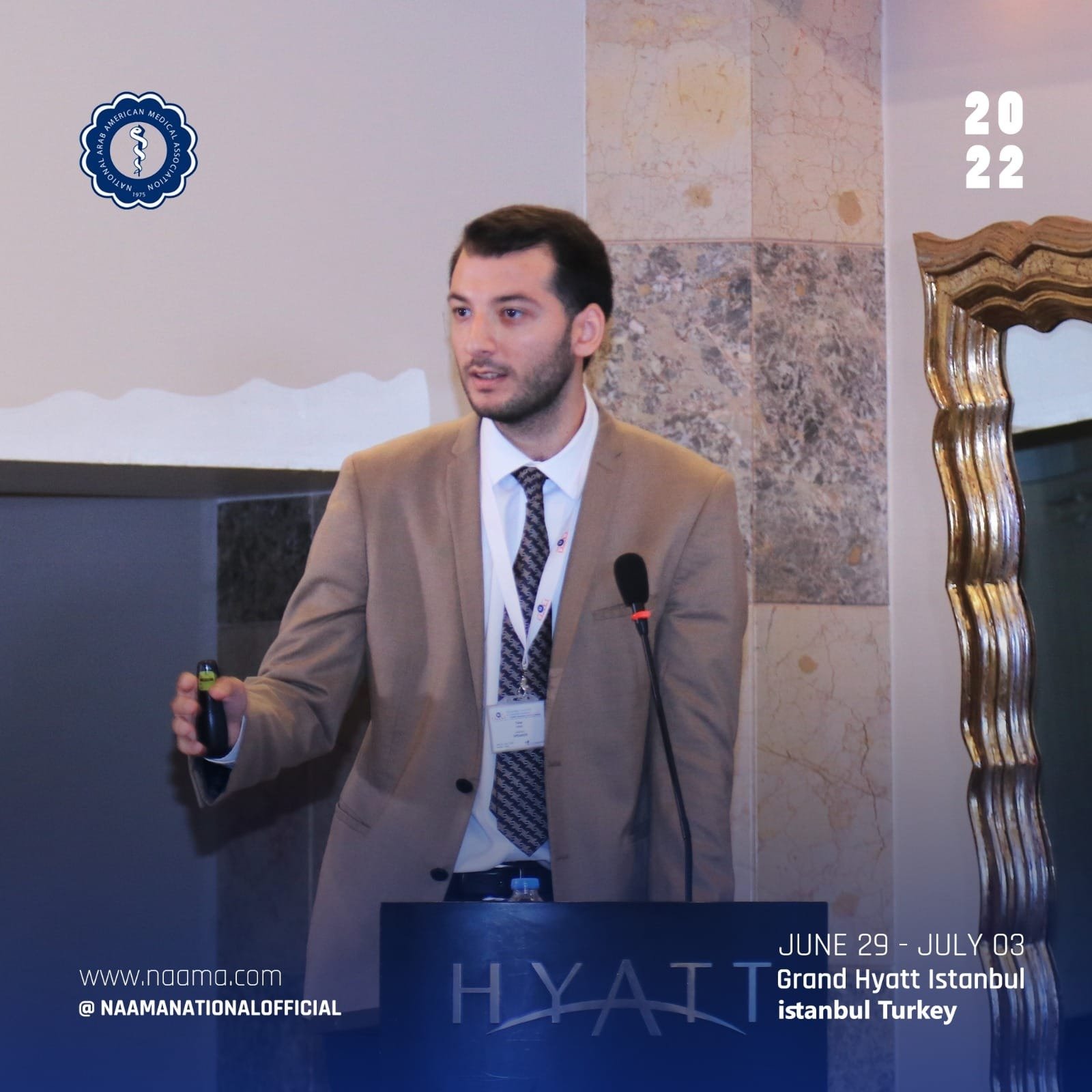 A man in a beige suit and tie giving a presentation at a conference, with a black microphone in front of him. There is a blue banner at the bottom indicating the event is from June 29 to July 3 at the Grand Hyatt Istanbul in Istanbul, Turkey. The bac