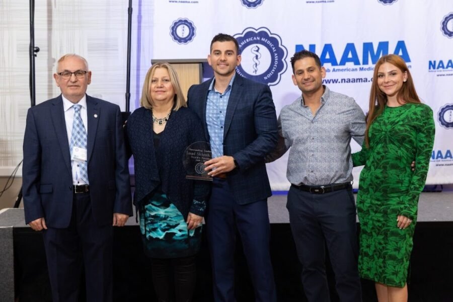 Group of five people at an award ceremony, standing in front of a backdrop with the NAAMA logo, with one person holding a glass award.