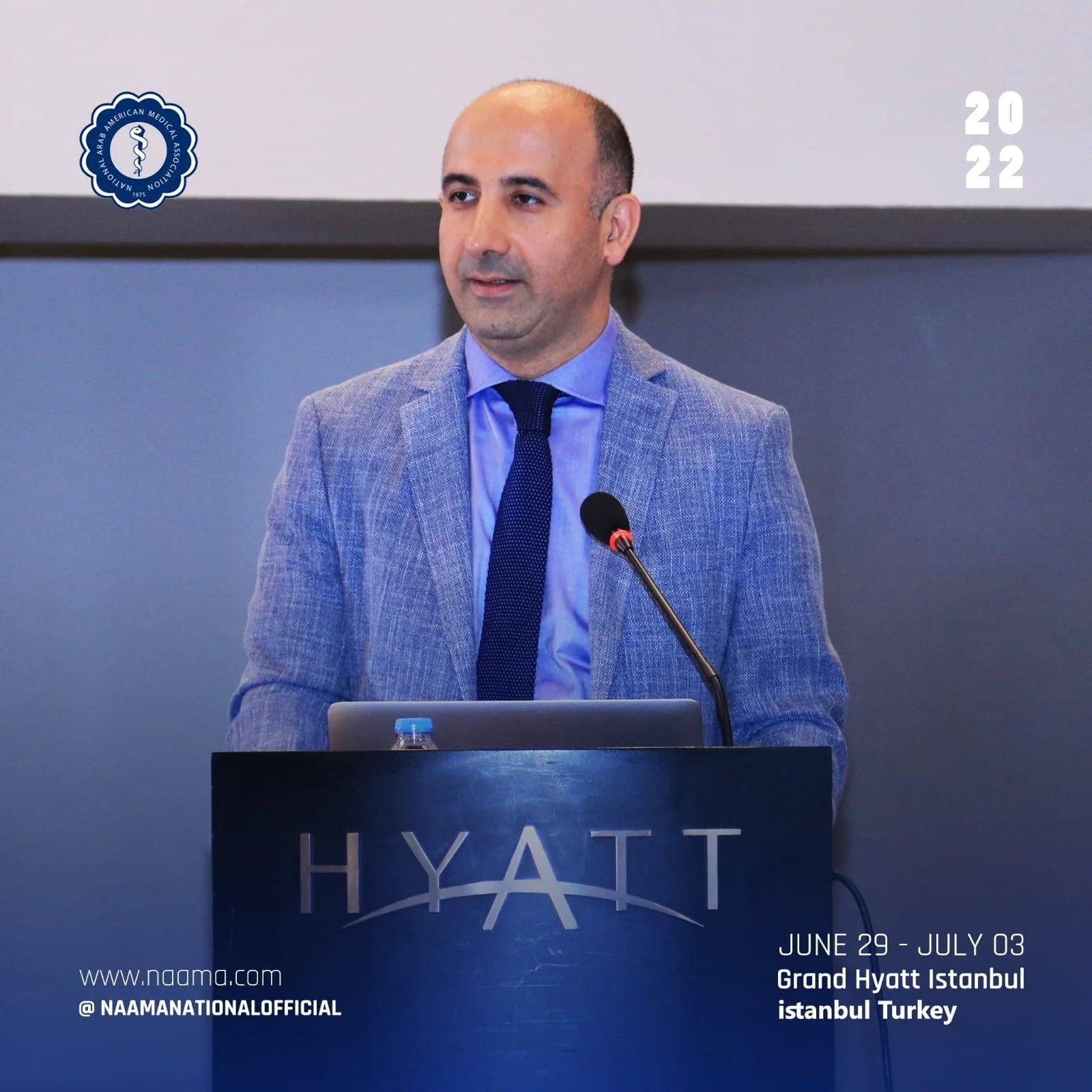A man in a gray blazer, blue shirt, and black tie speaking at a podium with a microphone. The background shows a sign for the Hyatt hotel and event details for June 29 to July 3 at the Grand Hyatt Istanbul in Turkey, along with the NAAM logo and webs