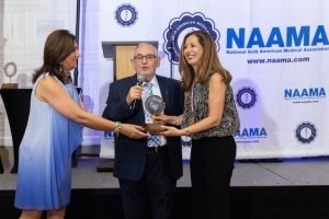 A woman in a blue dress presents an award to a woman in a black dress at a NAAMA event, with a man in a suit speaking into a microphone nearby.