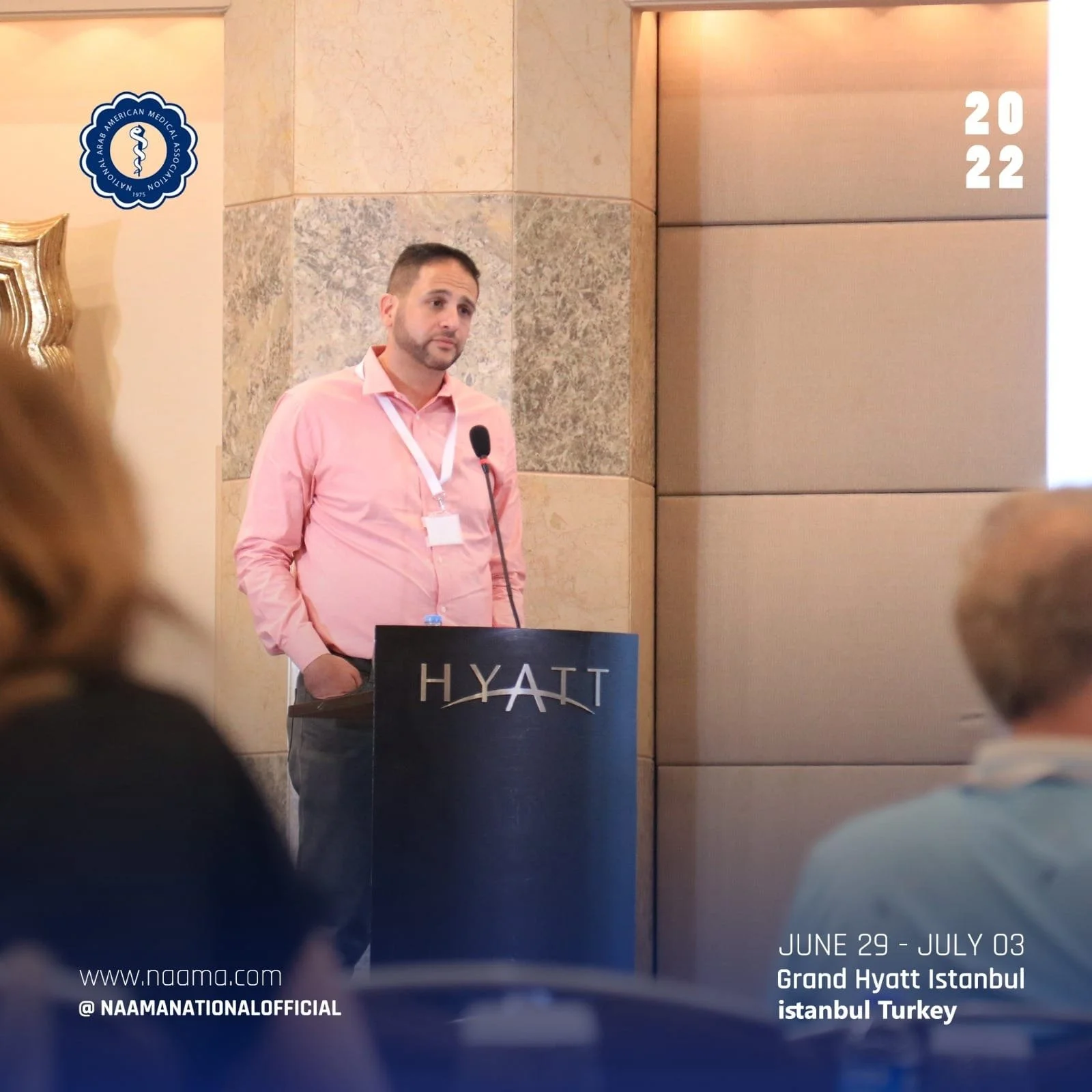 Man in pink shirt standing behind a podium at the Grand Hyatt Istanbul hotel, with a microphone, during an event from June 29 to July 3, 2022, organized by NAAM. The logo of the Arab American Medical Association is visible on the wall.