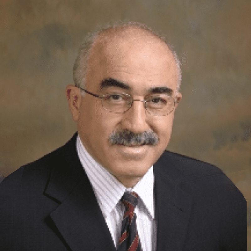 A middle-aged man with glasses, a mustache, and a bald head, wearing a dark suit, white striped shirt, and patterned tie, smiling at the camera.