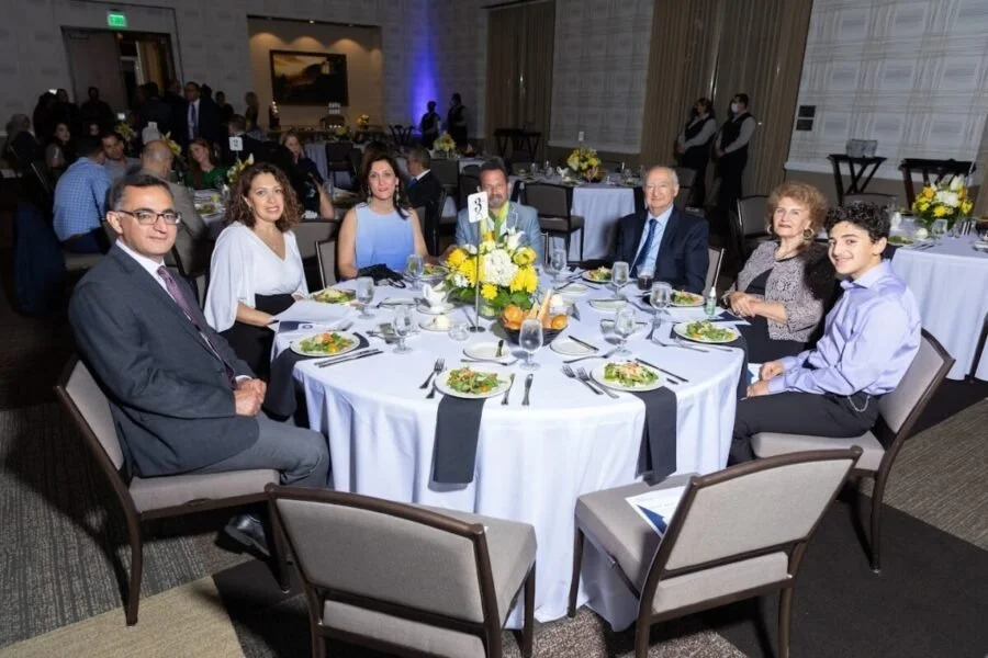 Group of eight people sitting around a round banquet table at a formal event with floral centerpiece, plates of salad, wine glasses, and silverware.