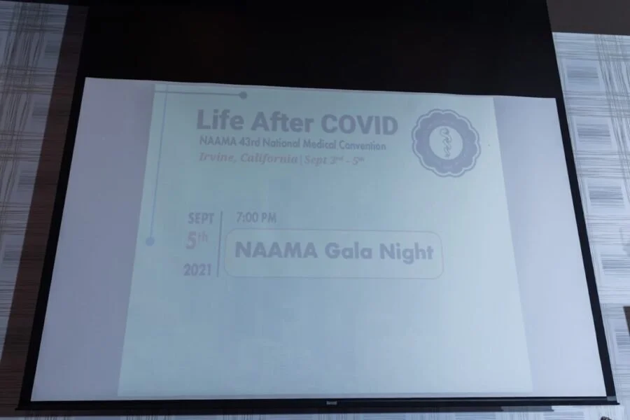 Projection of a presentation slide for a medical conference titled "Life After COVID" at NAAMA 43rd National Medical Convention in Guila, California, scheduled for September 5th, 2021, at 7:00 PM, with a logo of a caduceus symbol.