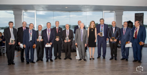 Group of 13 professionals in business attire posing together indoors with windows and blue sky in background.