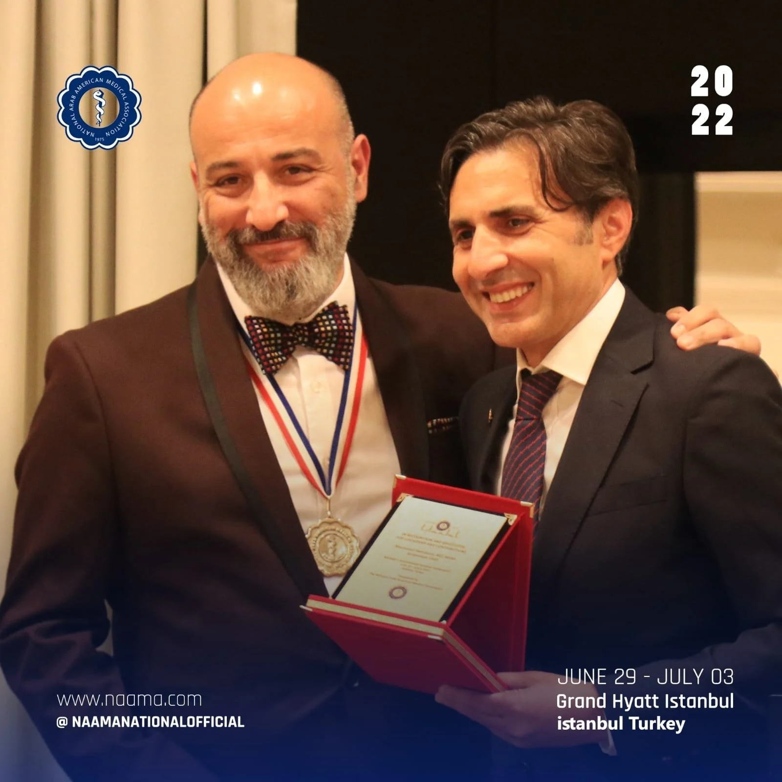Two men in formal attire smiling for a photo. The man on the left has a beard and is wearing a bow tie and an award medal around his neck. The man on the right has dark hair, is holding an award certificate, and has his arm around the other man. The 