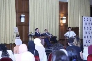 panel discussion with three men seated at a table on a stage, audience seated facing them, a man standing to the right speaking, in a room with yellow curtains and room lighting