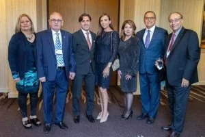 Group of seven people dressed in formal attire standing in a room with wood-paneled walls.