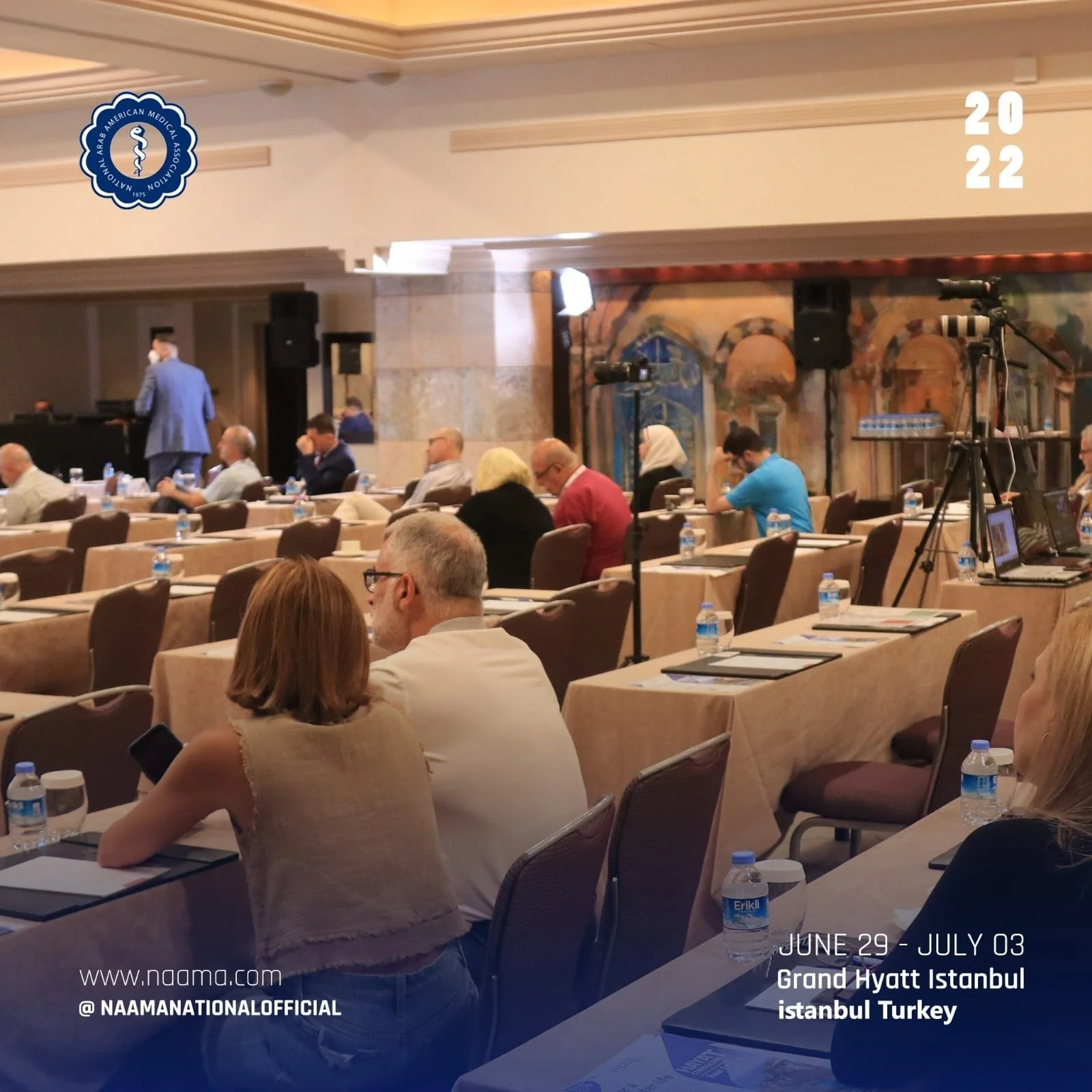 A conference room with attendees seated at tables, some wearing face masks. There are cameras and tripods set up, and bottled water on the tables. The room has artistic murals and a large logo of the Arab American Medical Association. The event is fr
