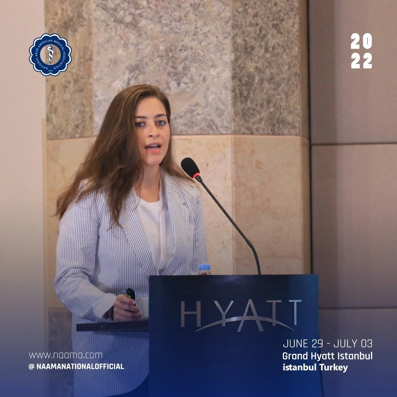 A woman with long brown hair speaking at a podium with the Hyatt logo, in a beige stone-tiled room at the Grand Hyatt Istanbul, Turkey, during an event from June 29 to July 3, 2022. The logo of the Arab American Medical Association is in the upper le